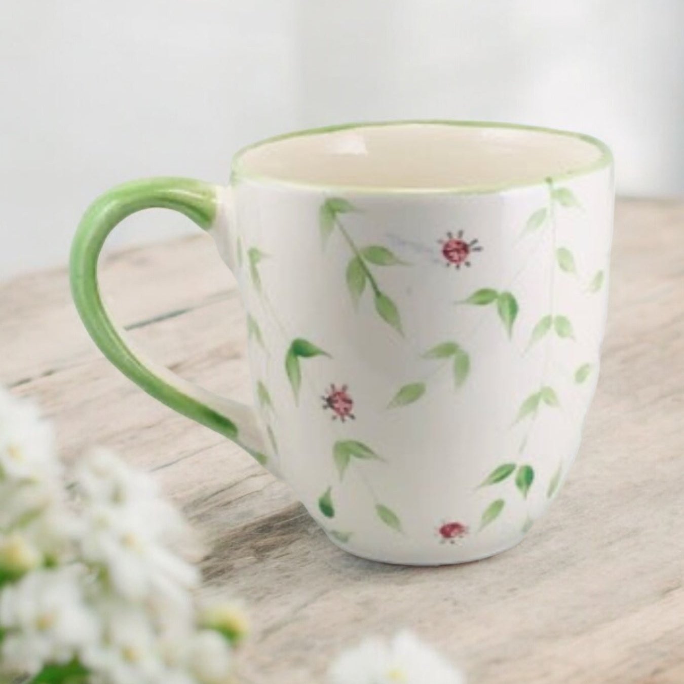 Hand painted ceramic extra large mug with floral green leaves with ladybugs patterns and a handle, displayed on a wooden surface with flowers in the background.