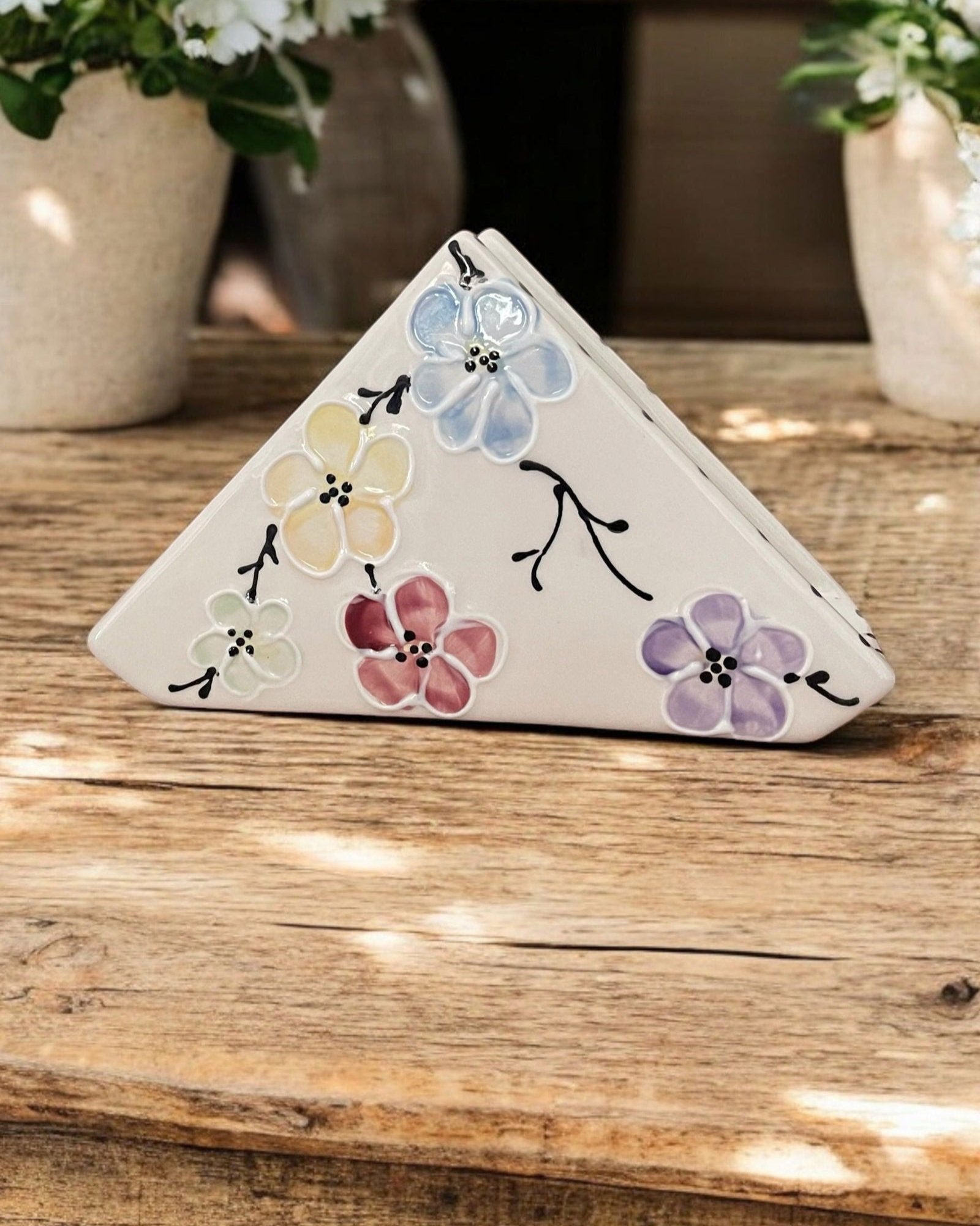 A triangular white ceramic napkin holder with painted colorful floral patterns displayed on a wooden table.
