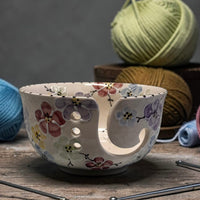 Ceramic yarn bowl with multi color cherry blossom floral design on a wooden surface with yarn and knitting needles in the background.