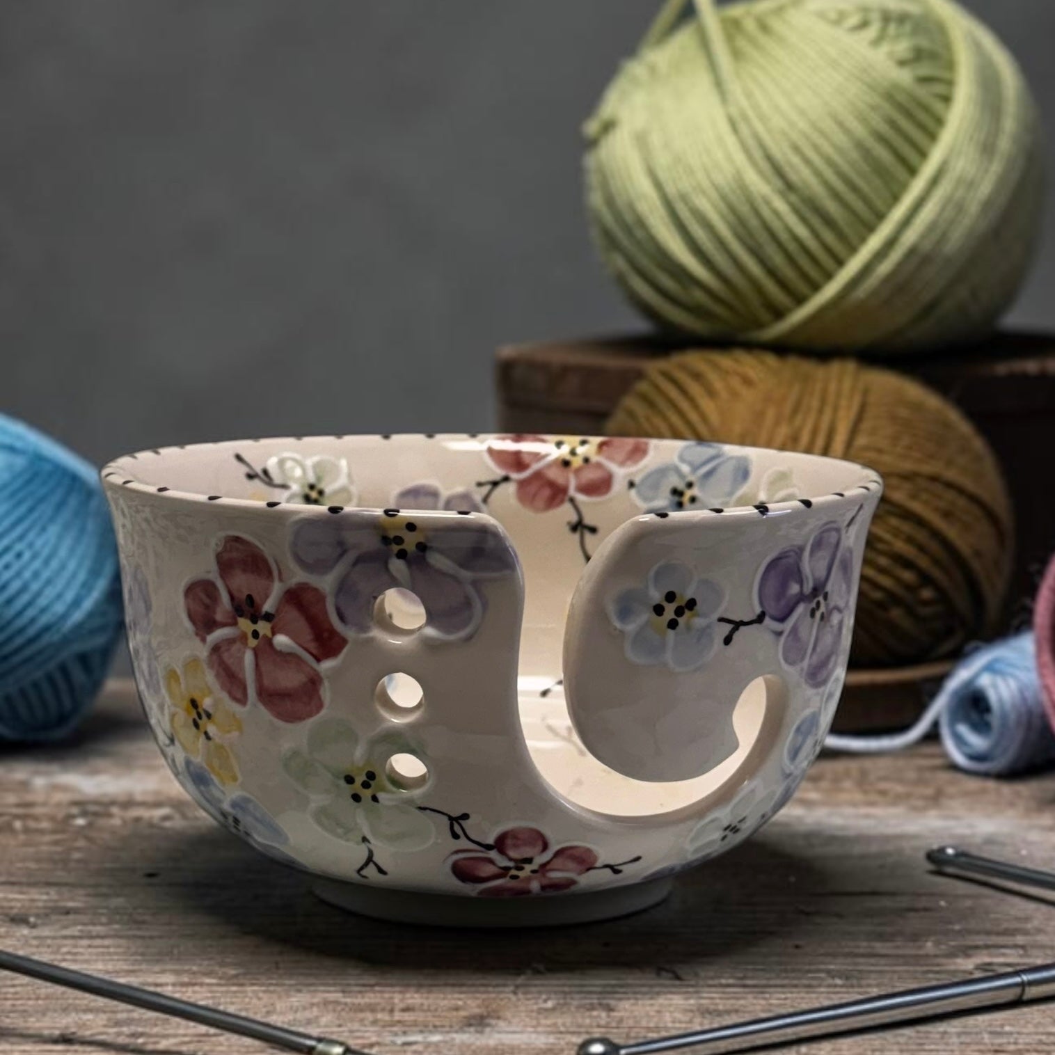 Ceramic yarn bowl with multi color cherry blossom floral design on a wooden surface with yarn and knitting needles in the background.