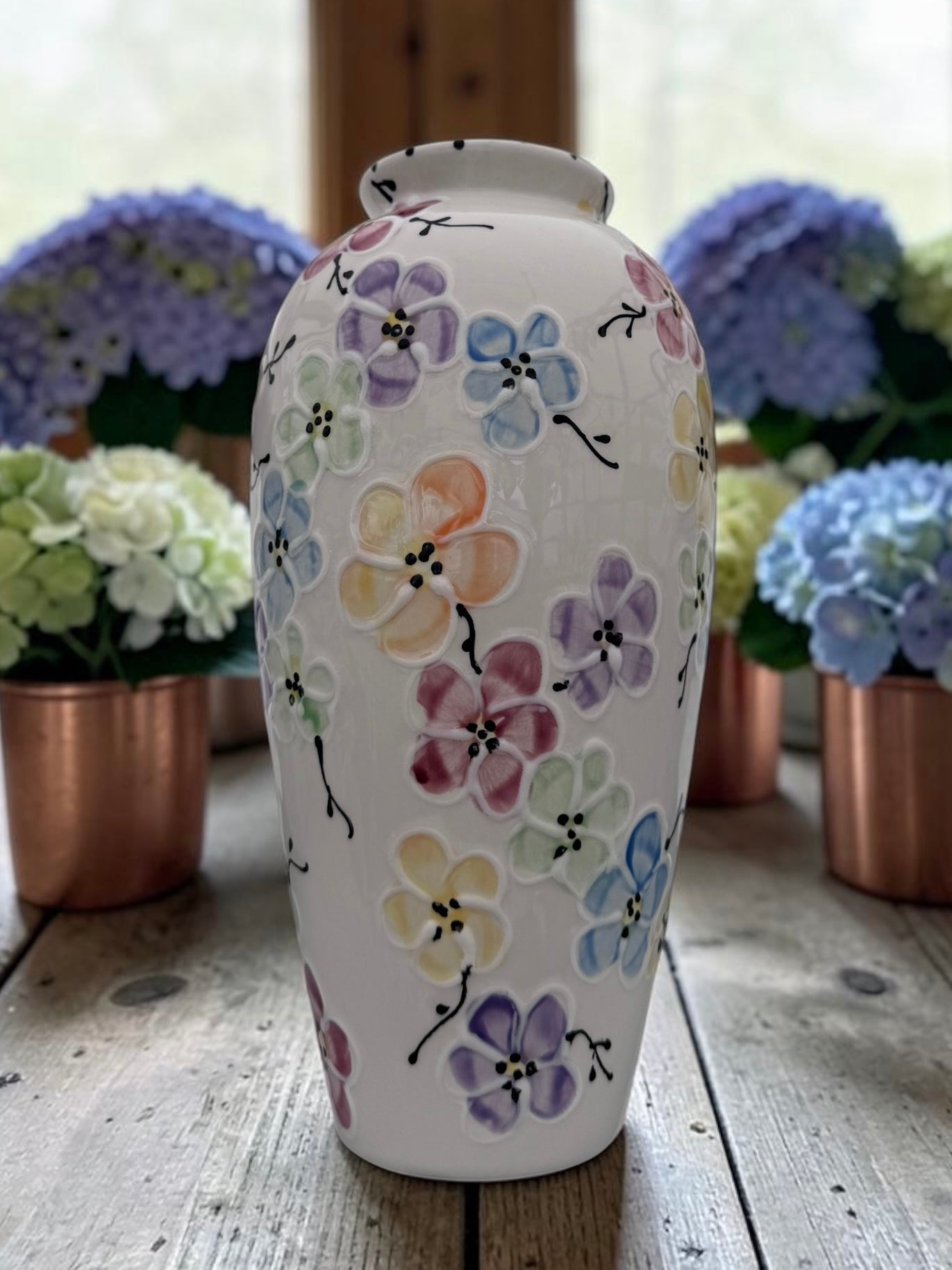 A white ceramic vase decorated with hand-painted pastel flowers in purple, pink, yellow, and blue, displayed on a rustic wooden table with blurred hydrangea arrangements in the background.