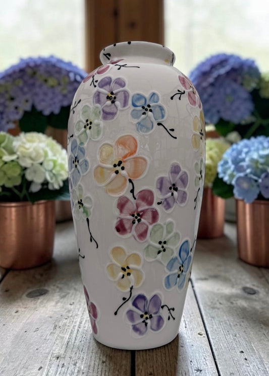 A white ceramic vase decorated with hand-painted pastel flowers in purple, pink, yellow, and blue, displayed on a rustic wooden table with blurred hydrangea arrangements in the background.