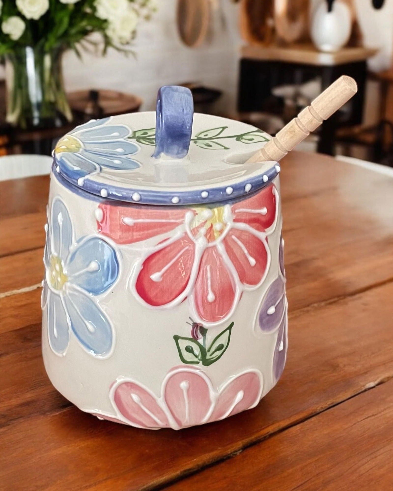 A ceramic honey jar with a lid, decorated with colorful hand-painted flowers in pink, blue, and purple on a cream background. It features a wooden spoon resting inside and is placed on a wooden table.