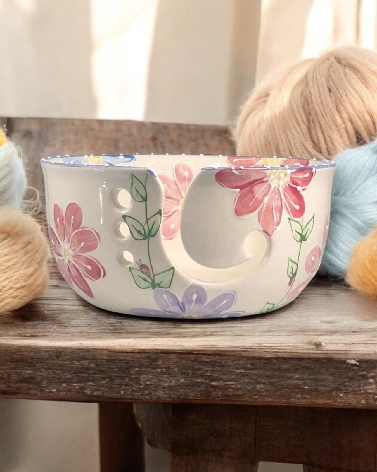 A hand-painted ceramic knitting bowl with floral painted design, containing yarn balls and knitting tools, placed on a wooden surface. Ladybird Ceramics Canada