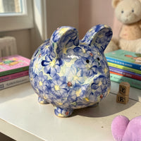 New Daisy Blue Floral-patterned piggy bank on a surface with books and toys in the background - Ladybird Ceramics