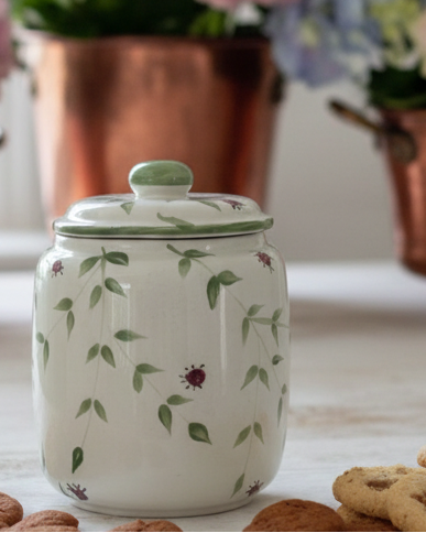 A white ceramic lidded jar decorated with green vines and small red ladybugs, set on a tabletop with cookies in the foreground and blurred copper pots of flowers in the background.
