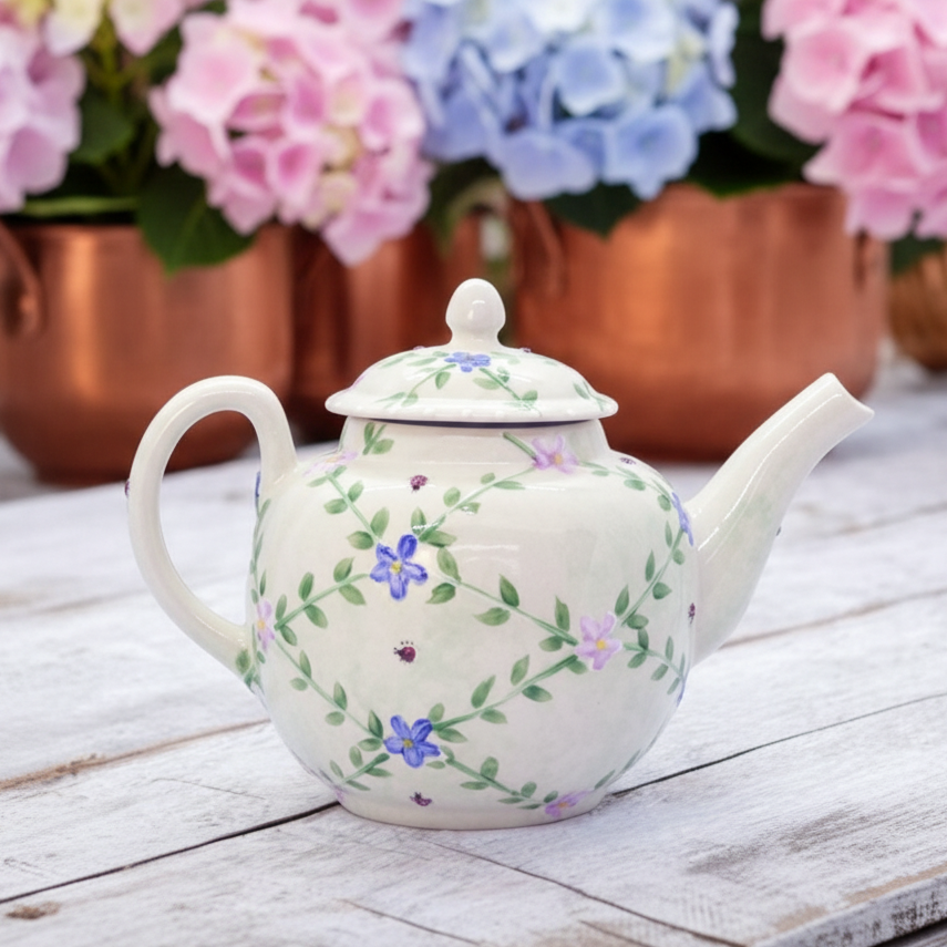 An off-white ceramic teapot decorated with a delicate green vine and purple/blue floral pattern, resting on a rustic white wooden table with blurred pink and blue hydrangeas in the background.