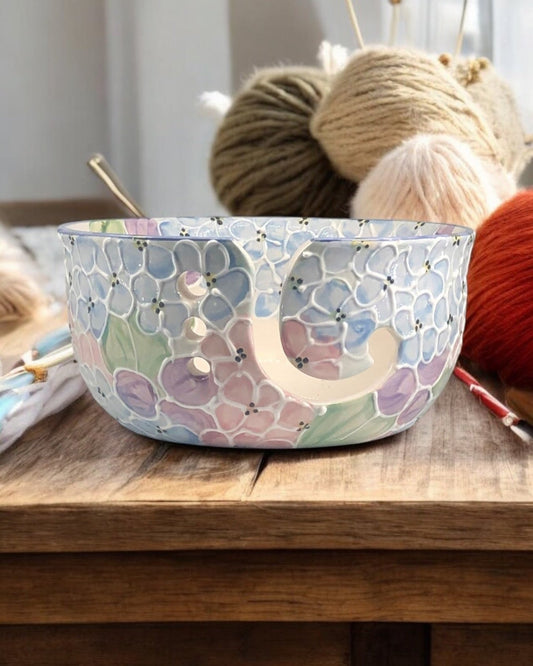 A hand-painted ceramic knitting bowl with hydrangea painted design, containing yarn balls and knitting tools, placed on a wooden surface. Ladybird Ceramics Canada