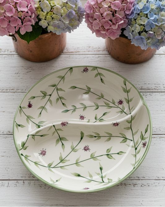 A ceramic two-piece serving platter with a green vine and purple flower pattern on an off-white background, styled on a white wooden table with copper pots of pink and blue hydrangeas in the background.
