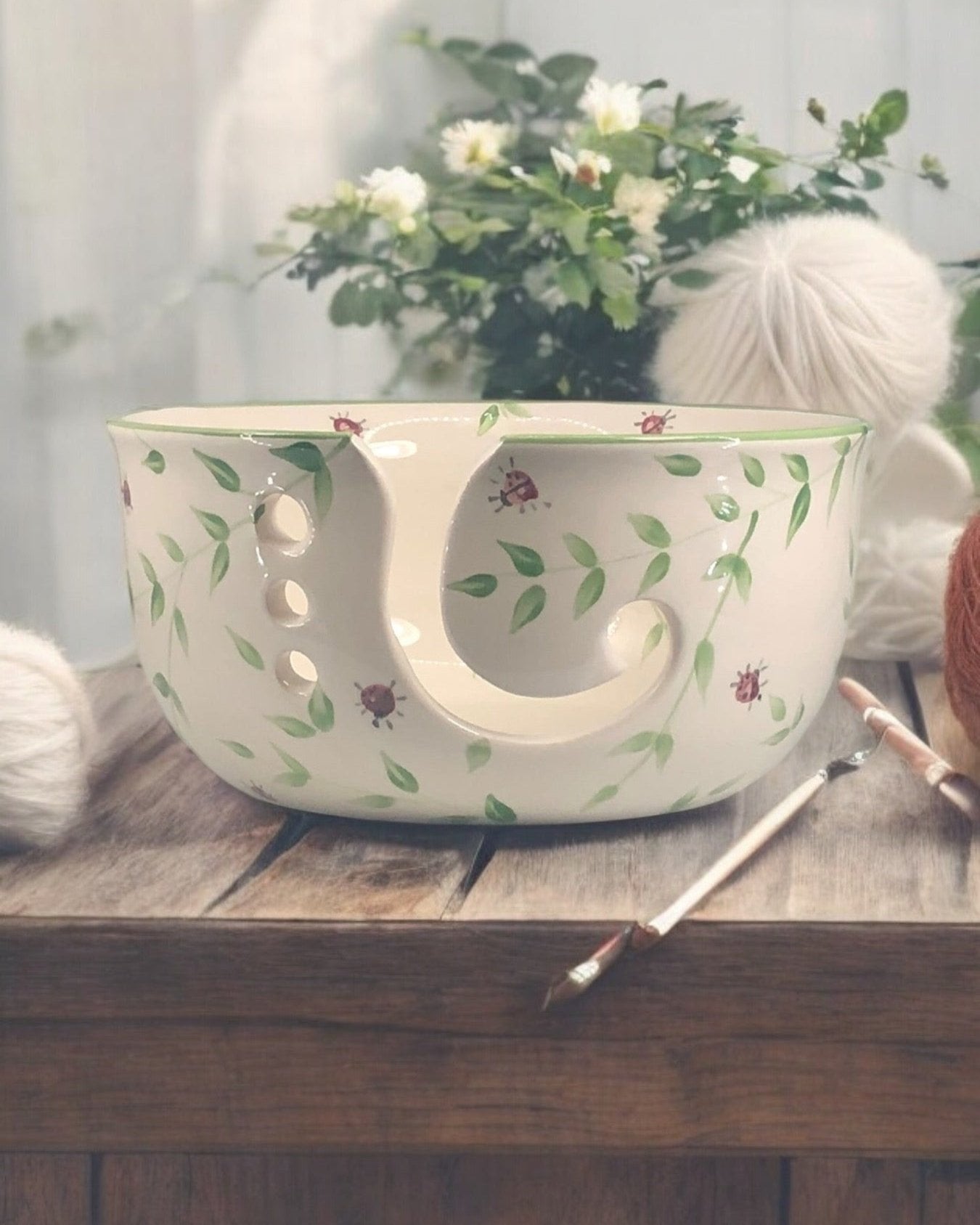 A hand-painted ceramic knitting bowl with leaves and ladybug design, containing yarn balls and knitting tools, placed on a wooden surface. Ladybird Ceramics Canada