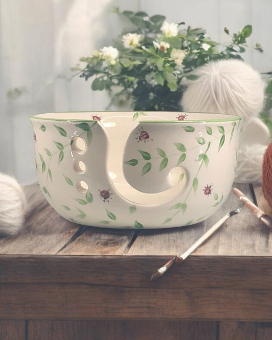 A hand-painted ceramic knitting bowl with leaves and ladybug design, containing yarn balls and knitting tools, placed on a wooden surface. Ladybird Ceramics Canada