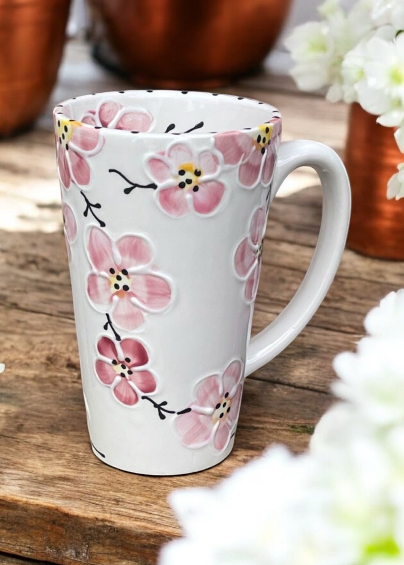A white ceramic mug decorated with pink cherry blossom flowers and black branches, set on a rustic wooden surface with blurred copper pots and white flowers in the background.