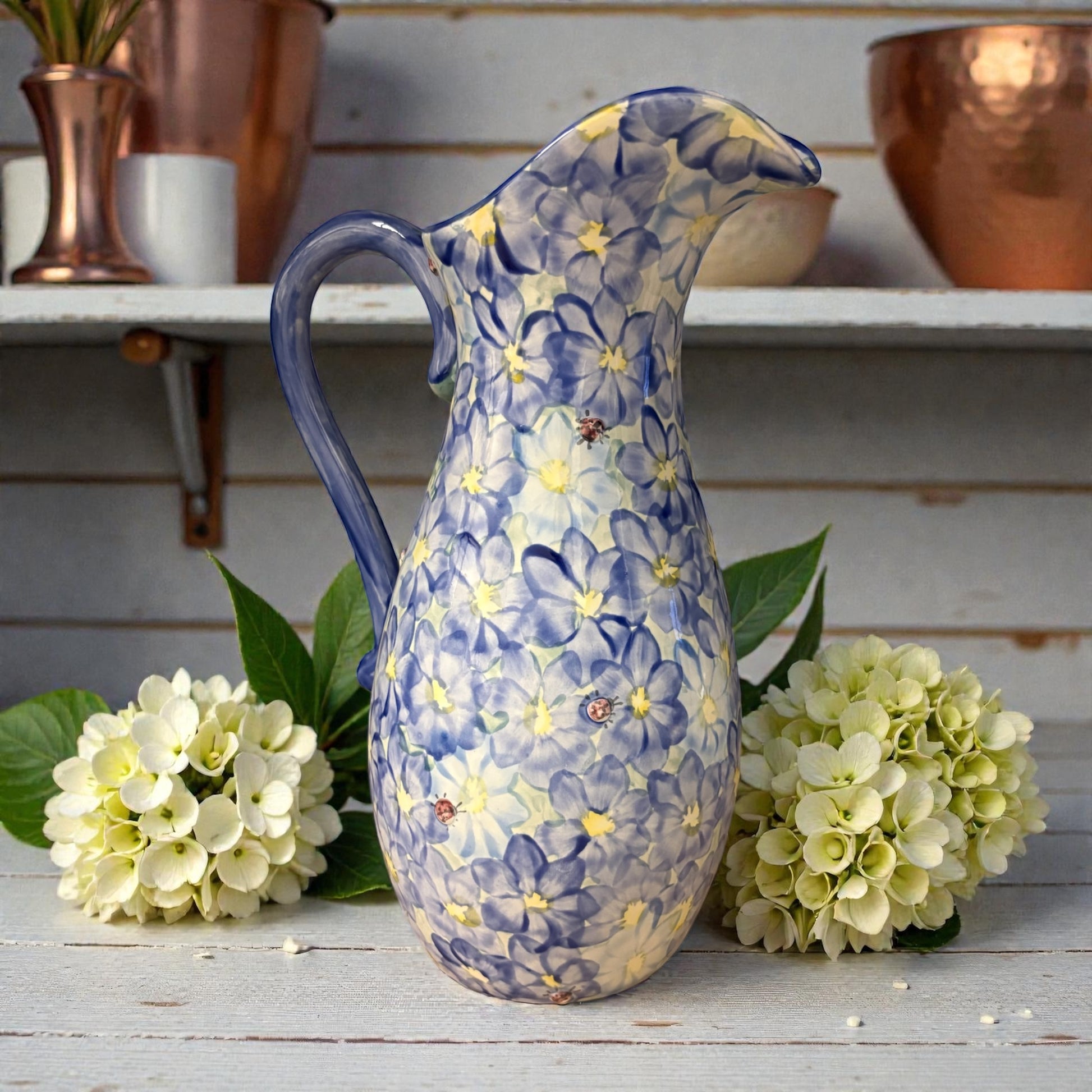 Decorative ceramic pitcher with daisy floral design on a wooden surface with flowers and shelves in the background.