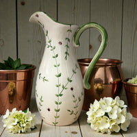 White ceramic elegant pitcher with green floral patterns on a wooden surface with copper pots and flowers. Ladybird Ceramics 