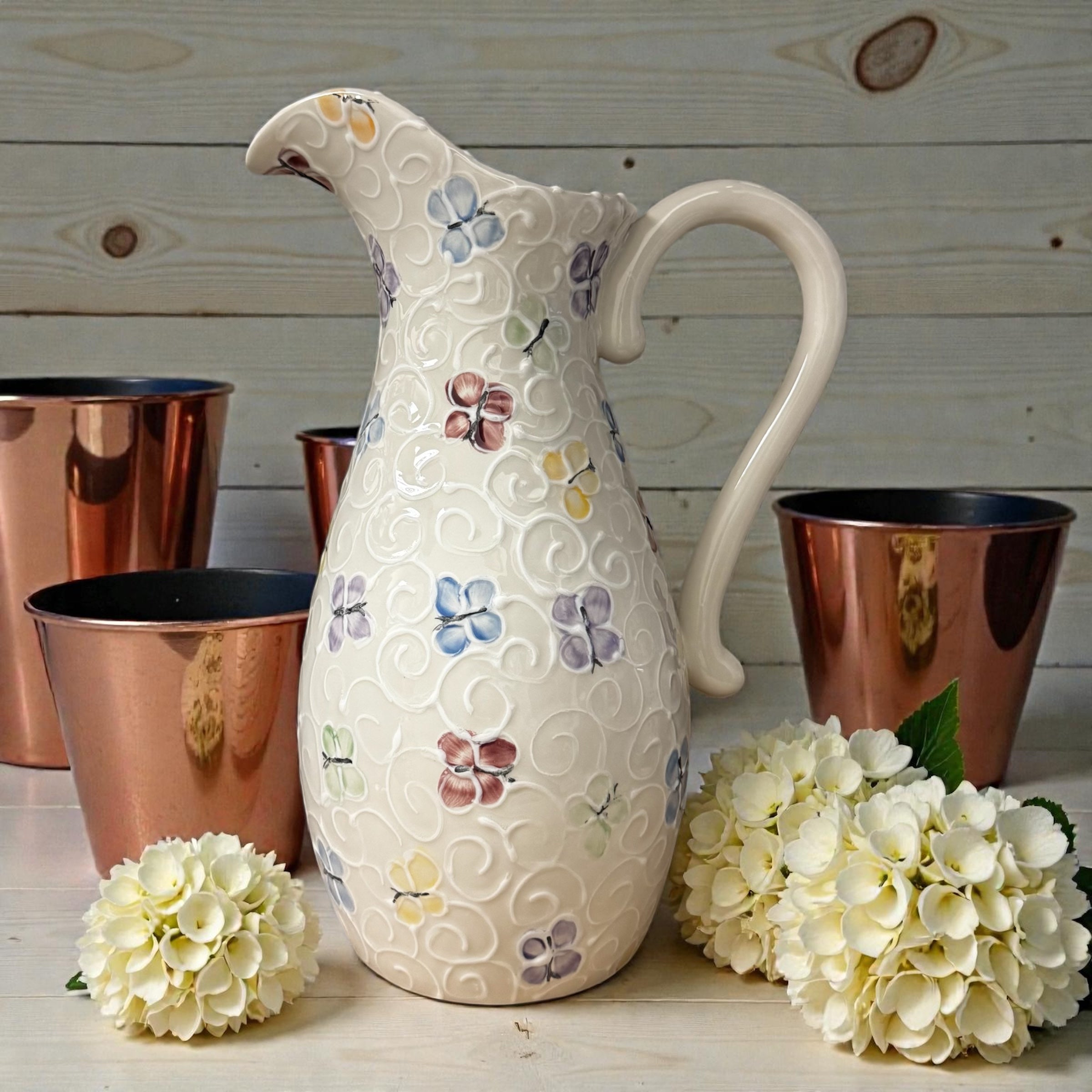 Decorative ceramic pitcher with butterfly patterns on a wooden surface with copper cups and flowers. Ladybird Ceramics
