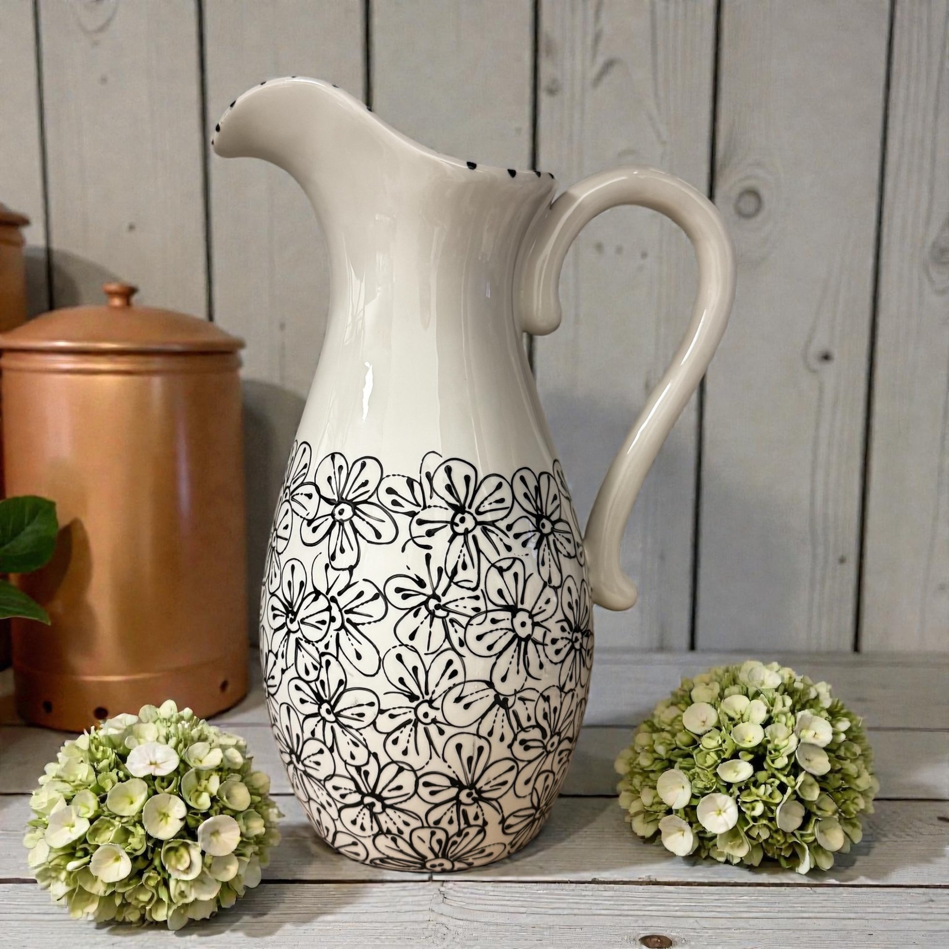 White ceramic pitcher with black floral pattern on a wooden surface with green flowers. Ladybird Ceramics
