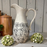 White ceramic pitcher with black floral pattern on a wooden surface with green flowers. Ladybird Ceramics