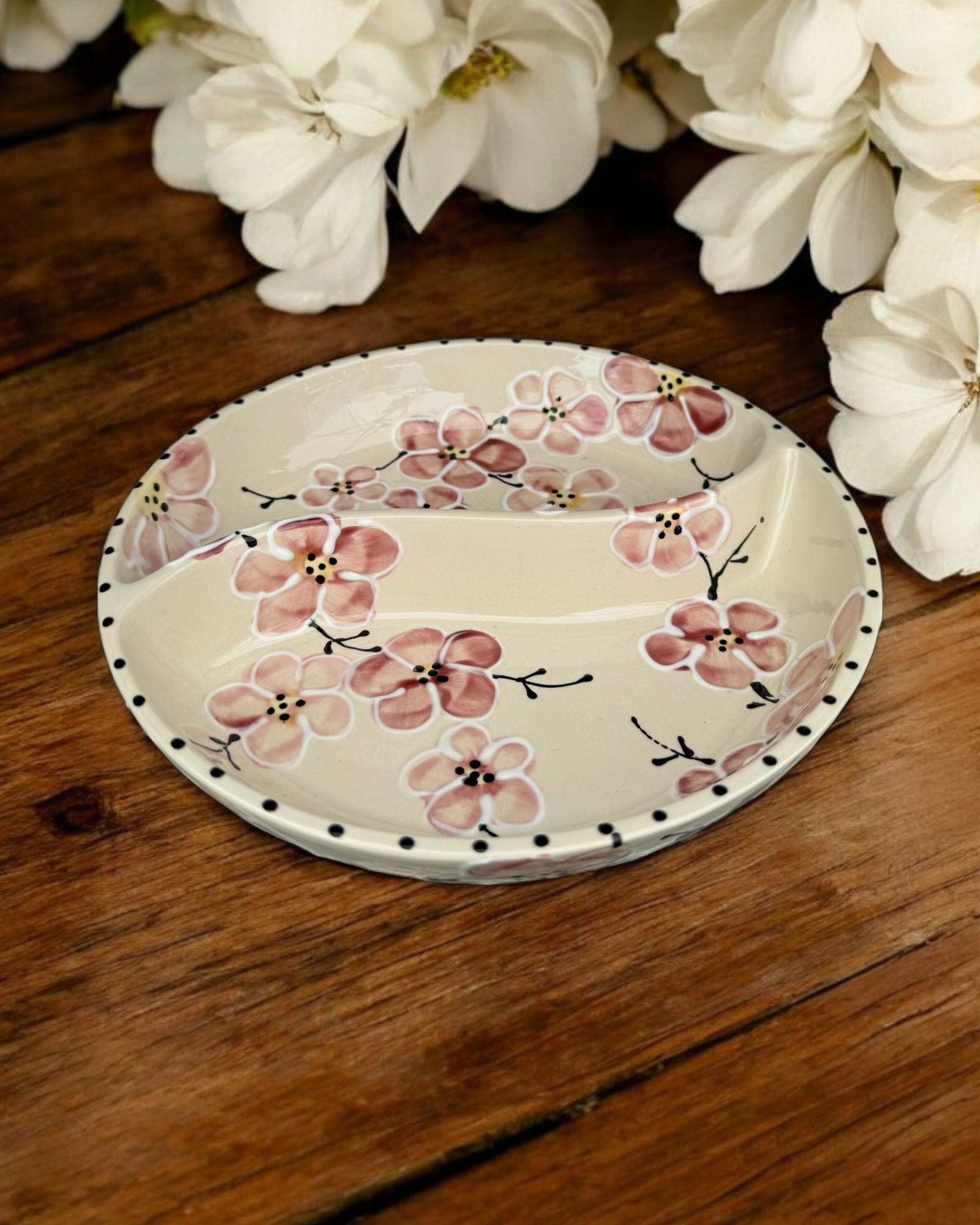A ceramic two-compartment serving Bowl with a white base decorated with a pink floral pattern, set on a white wooden surface with potted flowers in the background.