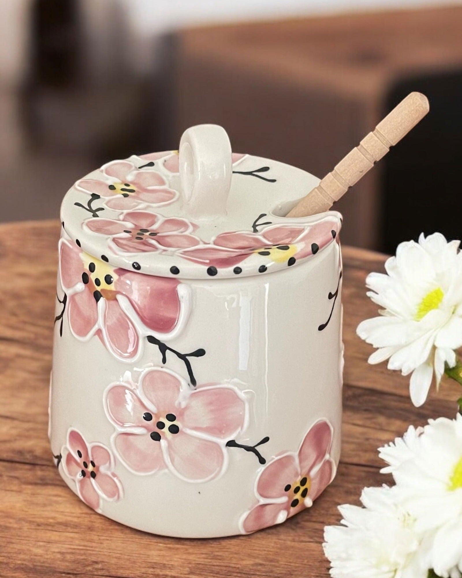 A white ceramic honey pot with pink cherry blossom designs, a wooden spoon, and white flowers on a wooden table.
