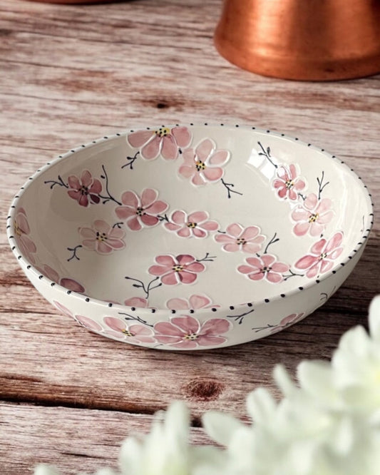 A hand-painted ceramic serving bowl with floral patterns, displayed on a wooden table with daisies in the foreground and copper cookware in the background.