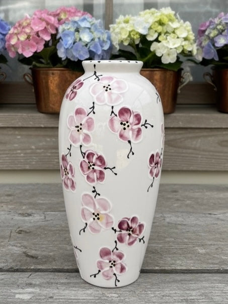 A white ceramic vase decorated with pink and purple cherry blossom flowers, standing on a weathered gray wooden surface with potted hydrangeas in the background.