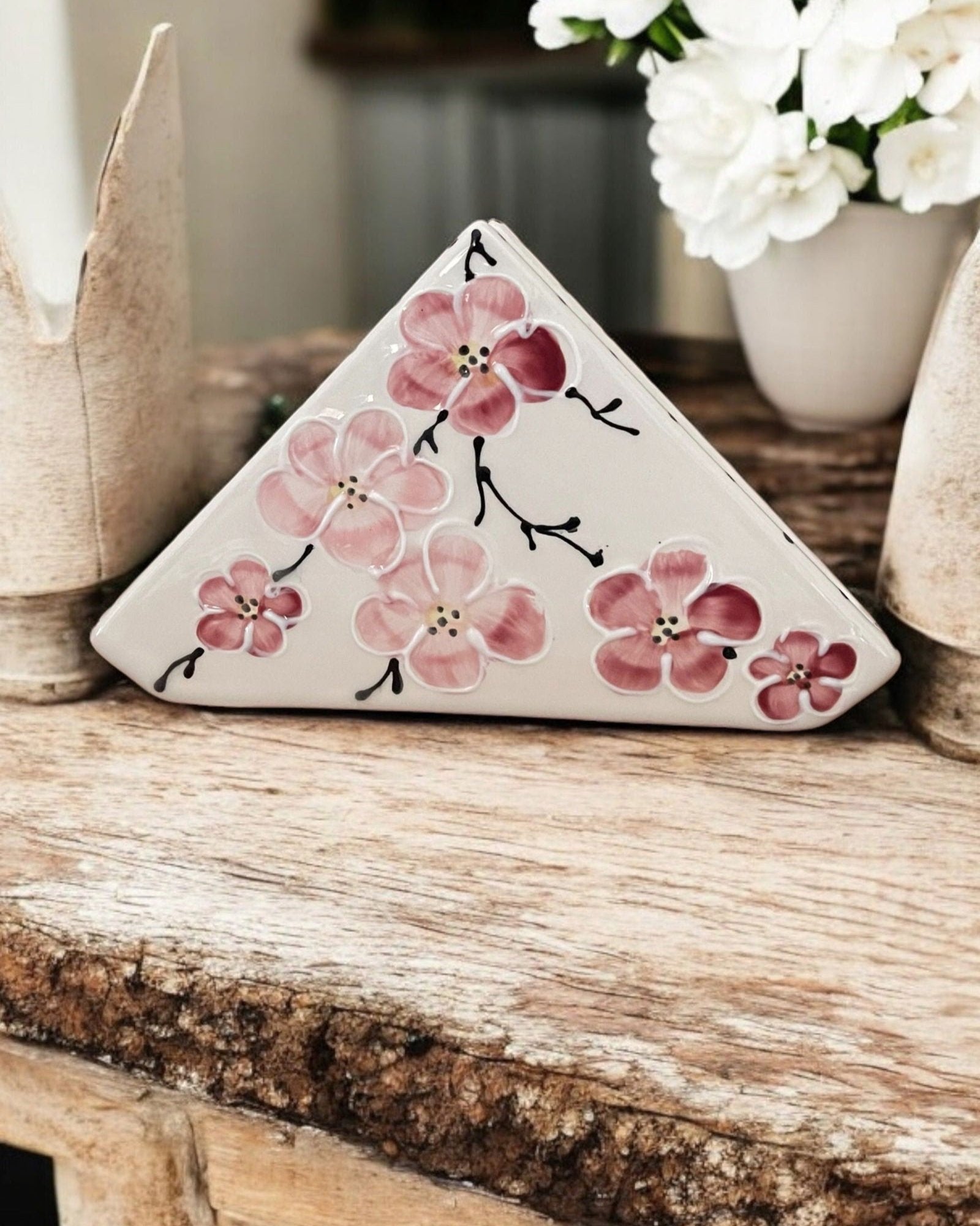 A triangular white ceramic napkin holder with painted pink floral patterns displayed on a wooden table.