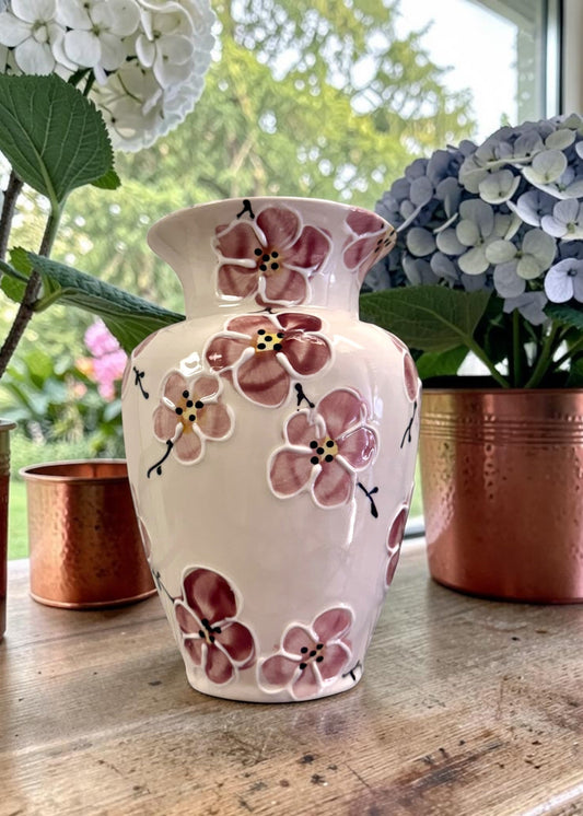 Ceramic vase with floral cherry blossom in pink design on a wooden surface with potted plants in the background