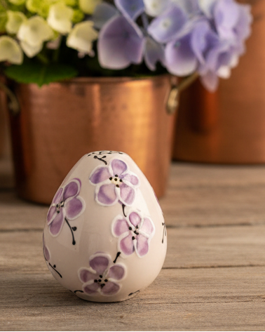 A small ceramic egg-shaped vase or keepsake box with a white glaze and hand-painted purple flowers, set on a rustic wooden surface with blurred copper planters and hydrangeas in the background.