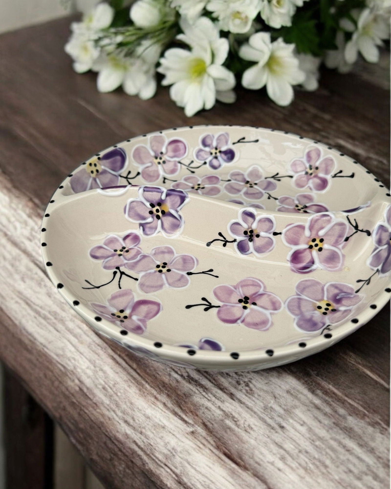 A ceramic two-compartment serving Bowl with a white base decorated with a colorful purple floral pattern, set on a white wooden surface with potted flowers in the background.