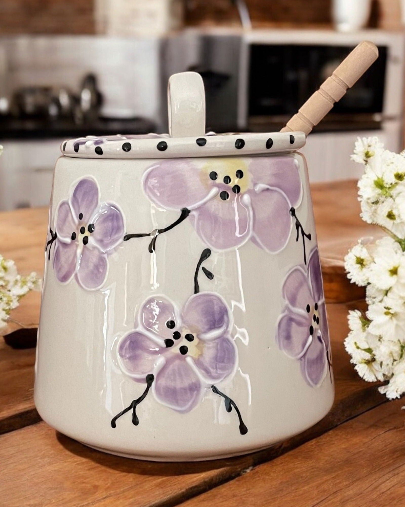 A white ceramic honey pot with pink cherry blossom designs, a wooden spoon, and white flowers on a wooden table.