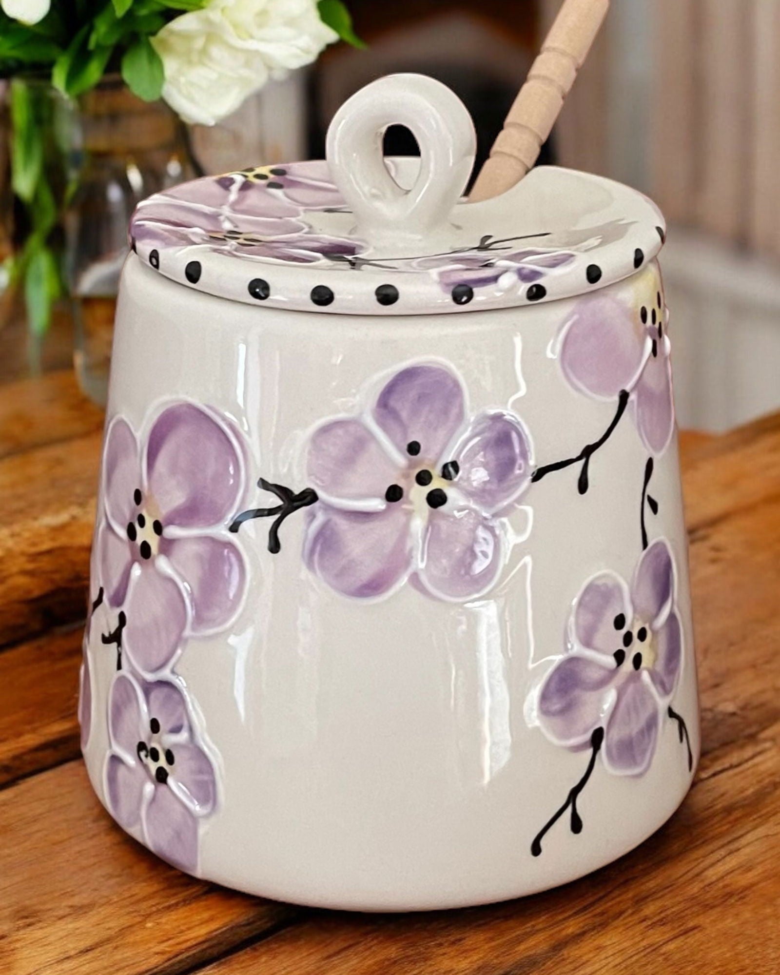 A cream-colored  ceramic honey pot jar with a lid and a wooden spoon, decorated with colorful floral motifs, set on a rustic wooden table. 