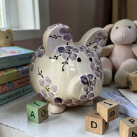 Decorative purple cherry blossom ceramic piggy bank with floral patterns on a surface with books and toys in the background - Ladybird Ceramics