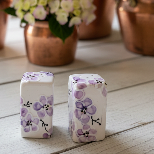 Two white ceramic salt and pepper shakers decorated with a purple floral pattern, set on a light wood table with blurred copper planters and flowers in the background.