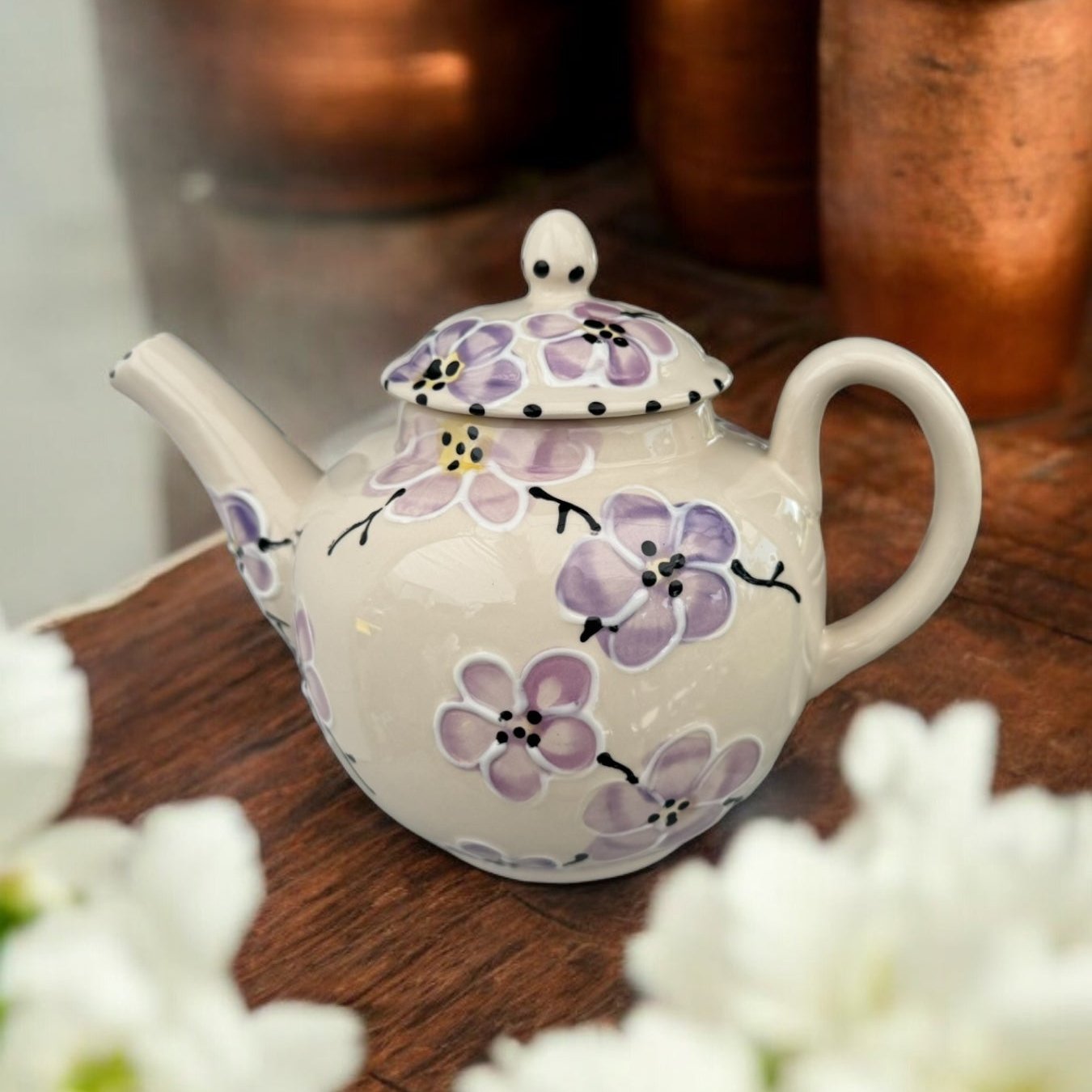 A white ceramic teapot with hand-painted floral designs, displayed on a wooden surface with a bouquet of white flowers beside it.