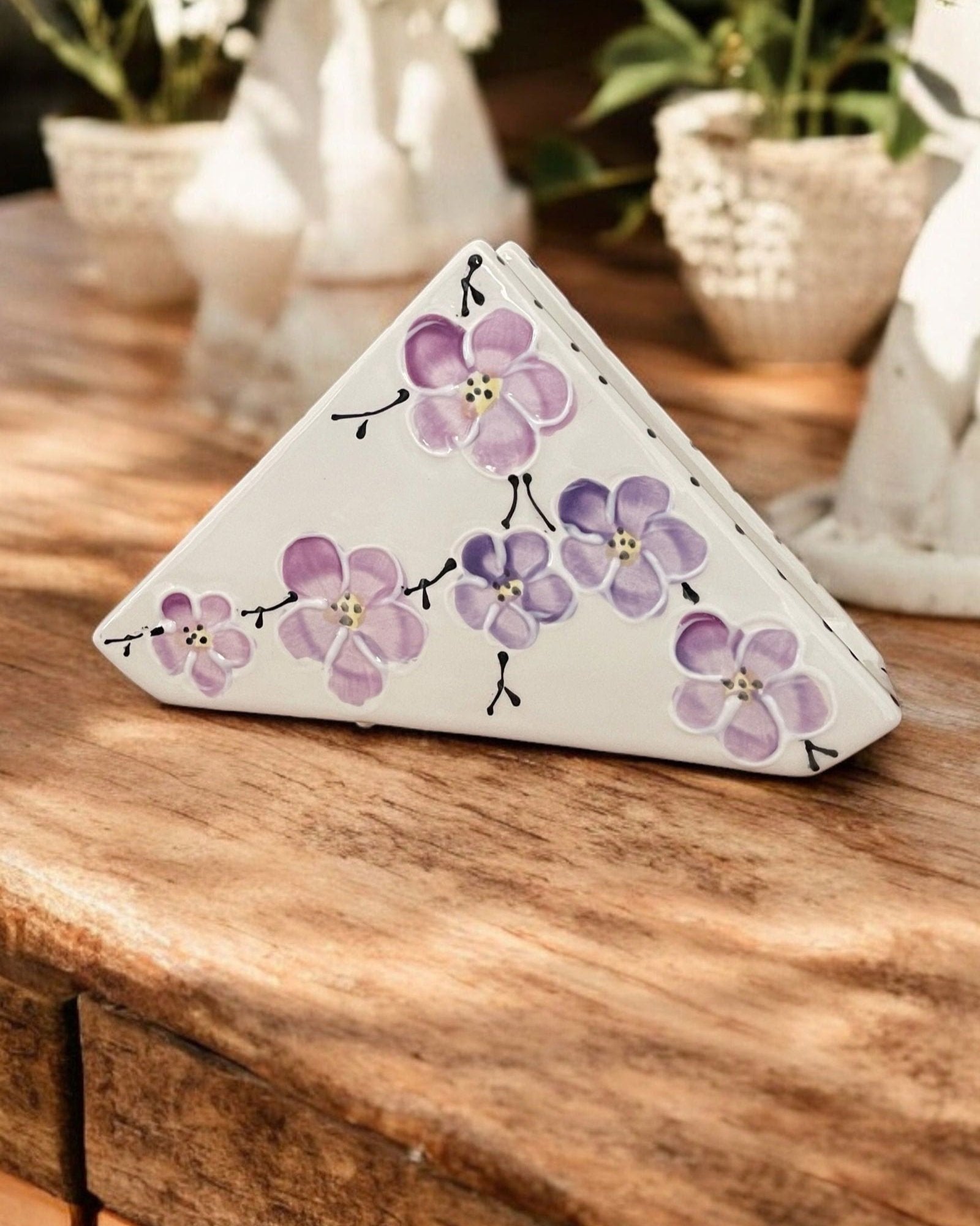 A triangular white ceramic napkin holder with painted purple floral patterns displayed on a wooden table.