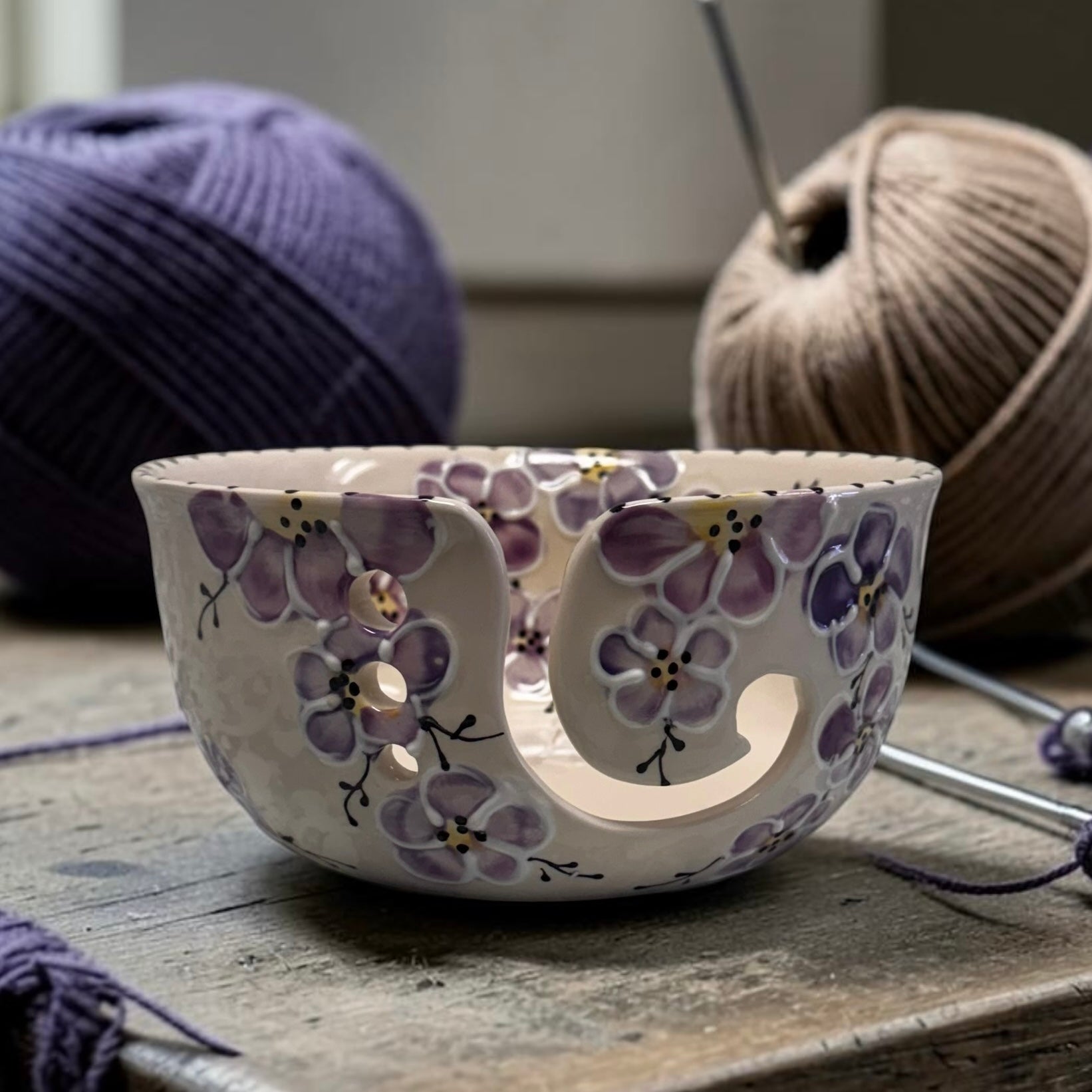 Ceramic yarn bowl with purple cherry blossom floral design on a wooden surface with yarn and knitting needles in the background. Ladybird Ceramics Canada
