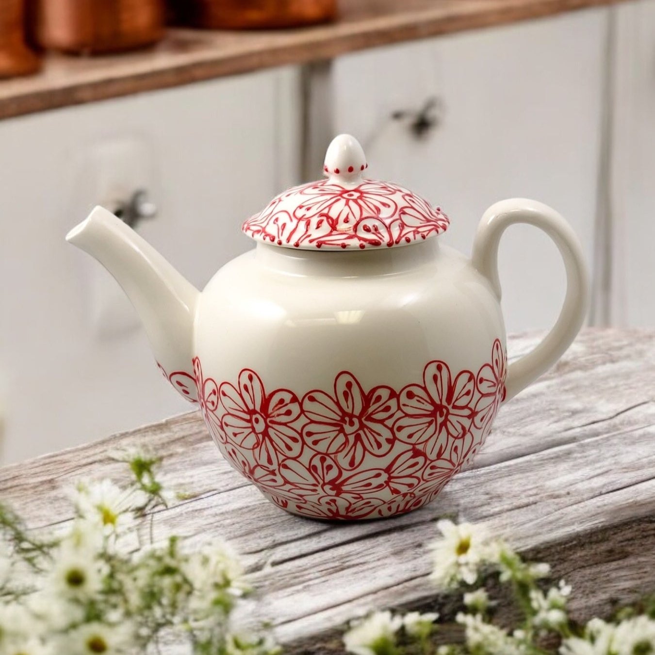 A white and red ceramic teapot with hand-painted floral designs, displayed on a wooden surface with a bouquet of white flowers beside it.