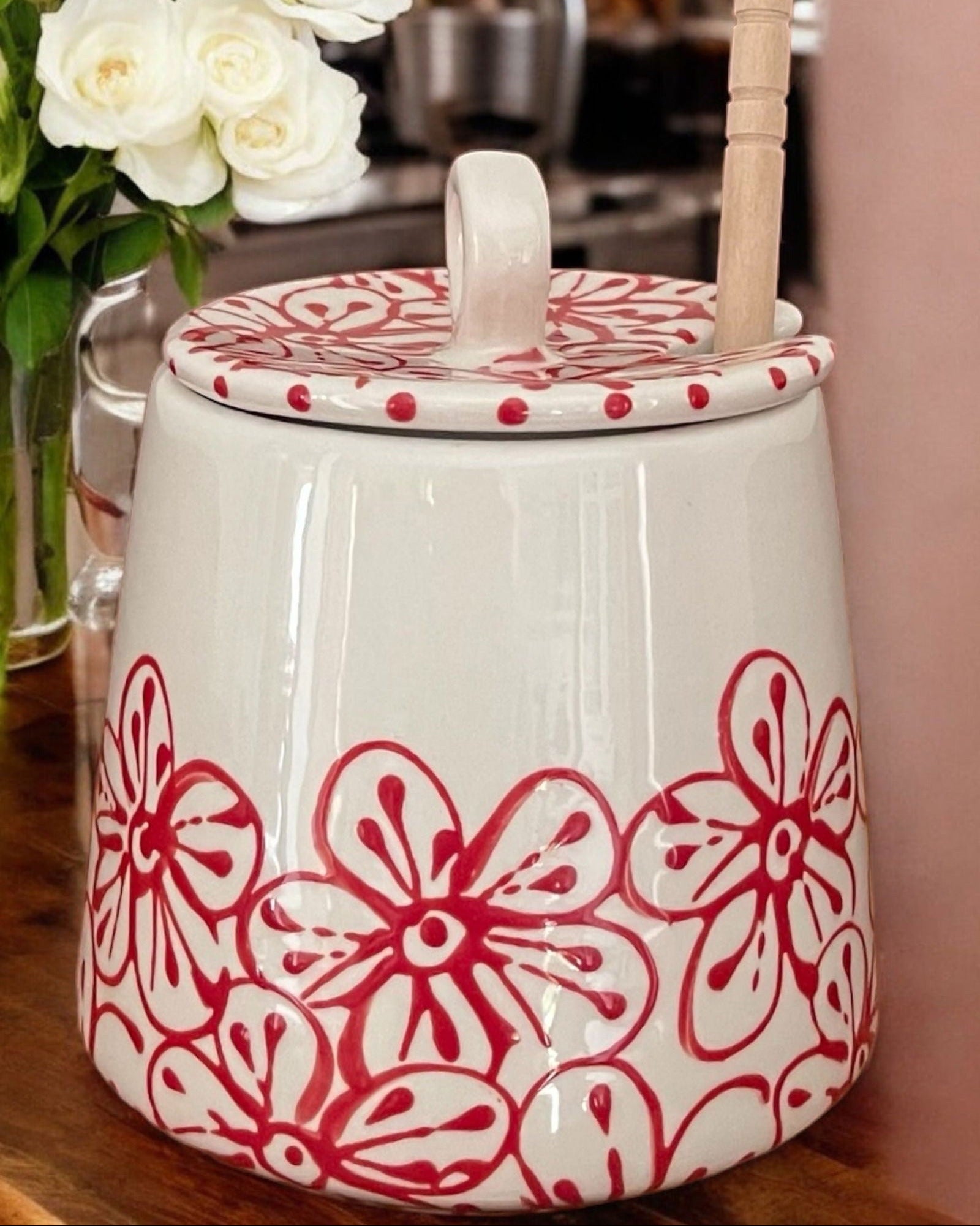 A white ceramic honey pot with a red blossom designs, a wooden spoon, and white flowers on a wooden table.