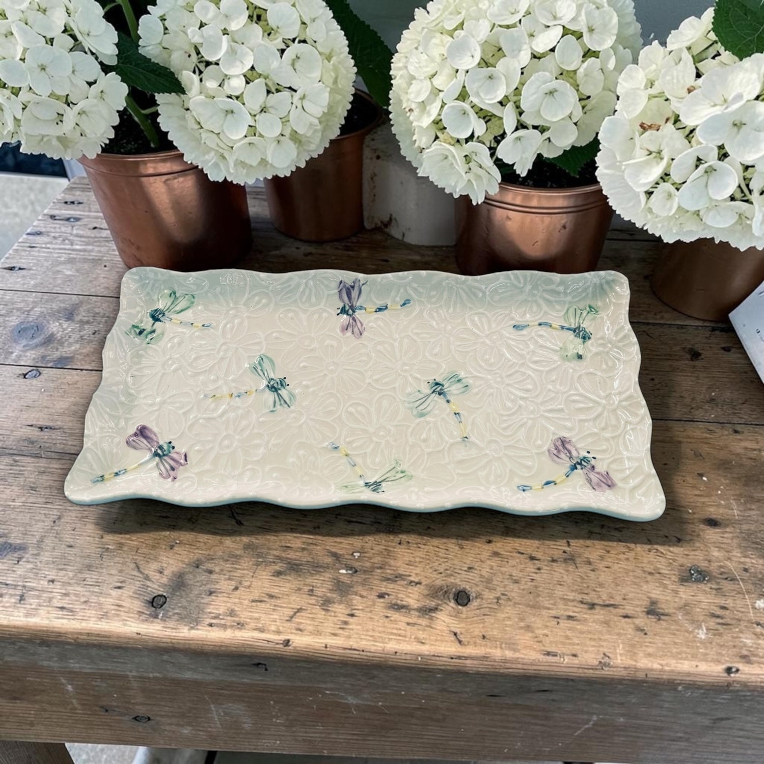 Decorative ruffle ceramic tray with white dragonfly patterns on a wooden surface with potted white flowers in the background.