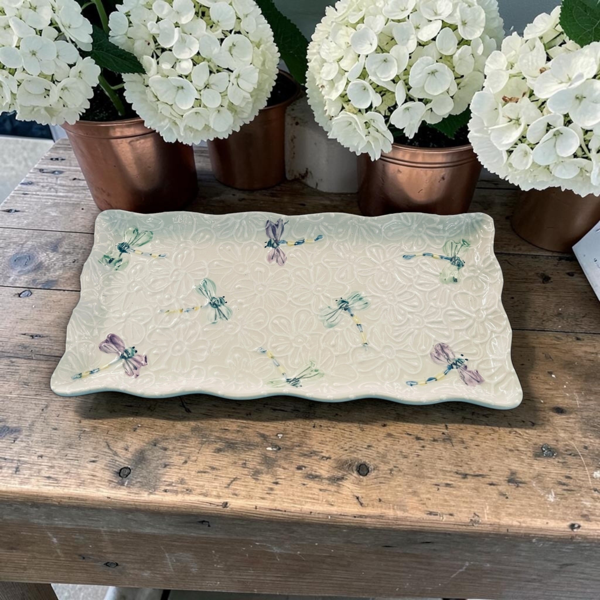 Decorative ruffle ceramic tray with white dragonfly patterns on a wooden surface with potted white flowers in the background.