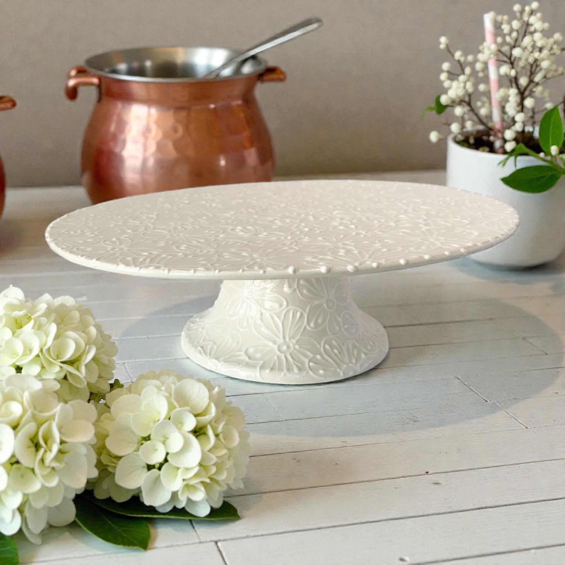 White decorative cake stand on a wooden surface with flowers and a pot in the background