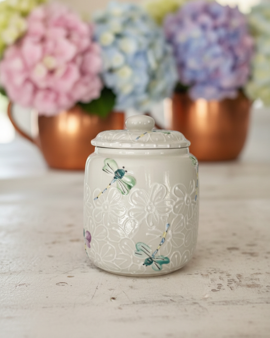 A small white ceramic jar with a lid, decorated with embossed floral patterns and painted green dragonflies, set on a light stone surface with blurred pink, blue, and purple hydrangeas in copper pots behind it.