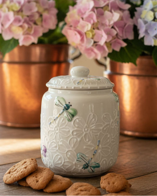 A white ceramic cookie jar with embossed floral patterns and painted dragonflies, styled on a wooden table with cookies in the foreground and copper pots of pink and blue hydrangeas in the background.