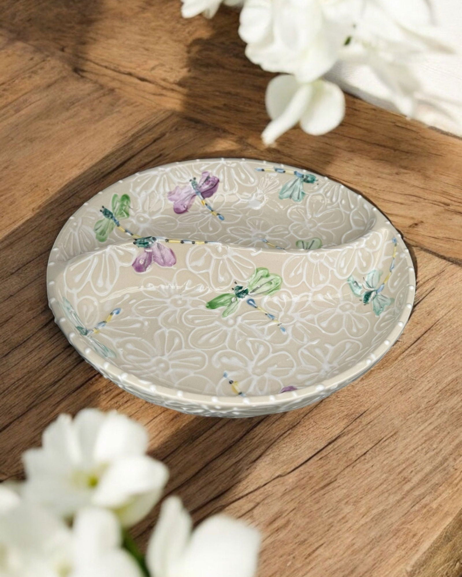 A ceramic two-compartment serving Bowl with a white base decorated with a dragonfly and floral pattern, set on a white wooden surface with potted flowers in the background.