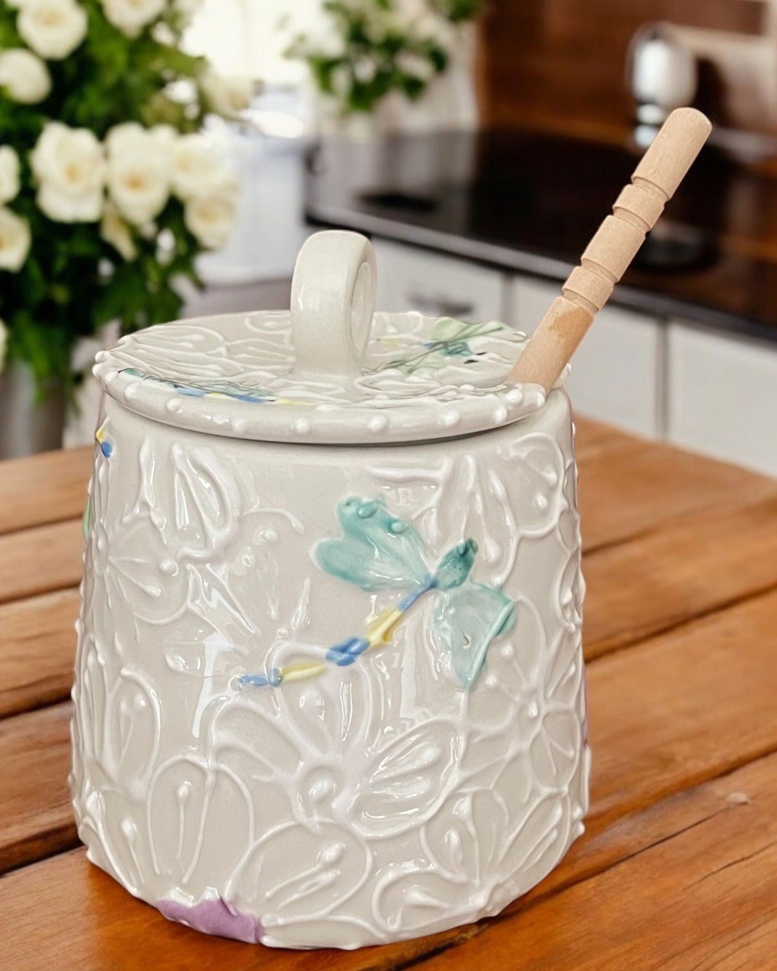 A white ceramic honey pot with embossed floral and dragonfly patterns, a matching lid with a handle, and a wooden dipper spoon, set on a wooden table with a blurred kitchen background.