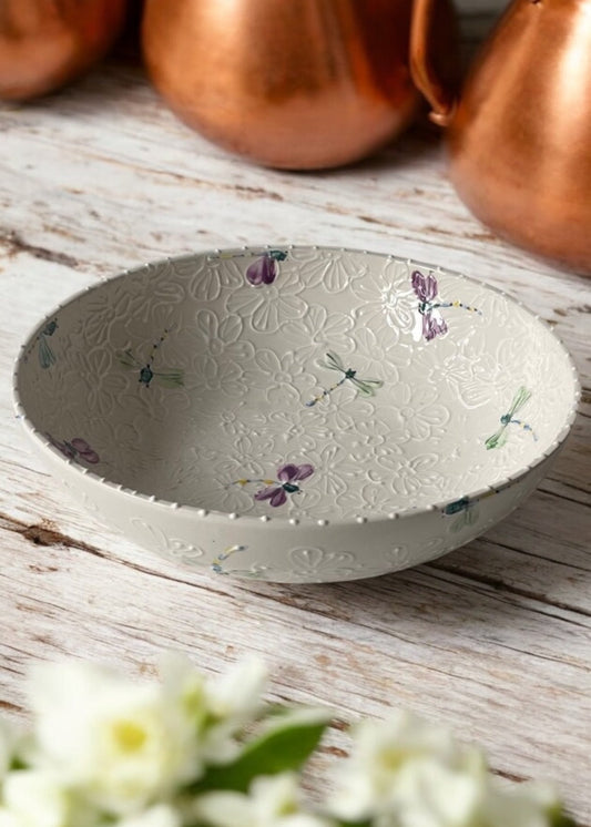 A hand-painted ceramic serving bowl with floral patterns, displayed on a wooden table with daisies in the foreground and copper cookware in the background.