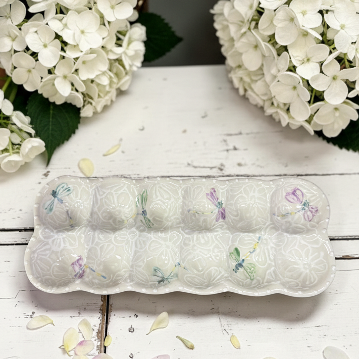A white ceramic egg tray with embossed floral patterns and small painted dragonflies in pastel colors, displayed on a white wooden surface with white flowers in the background.
