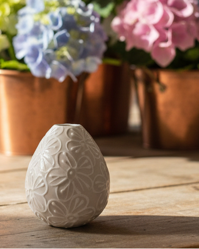 A small teardrop-shaped ceramic vase in a warm off-white color with an embossed floral pattern, sitting on a rustic wooden table with a blurred background of pink and blue hydrangeas in copper pots.