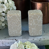 Two rectangular salt pepper ceramics  with floral patterns on a wooden surface with flowers in the background.