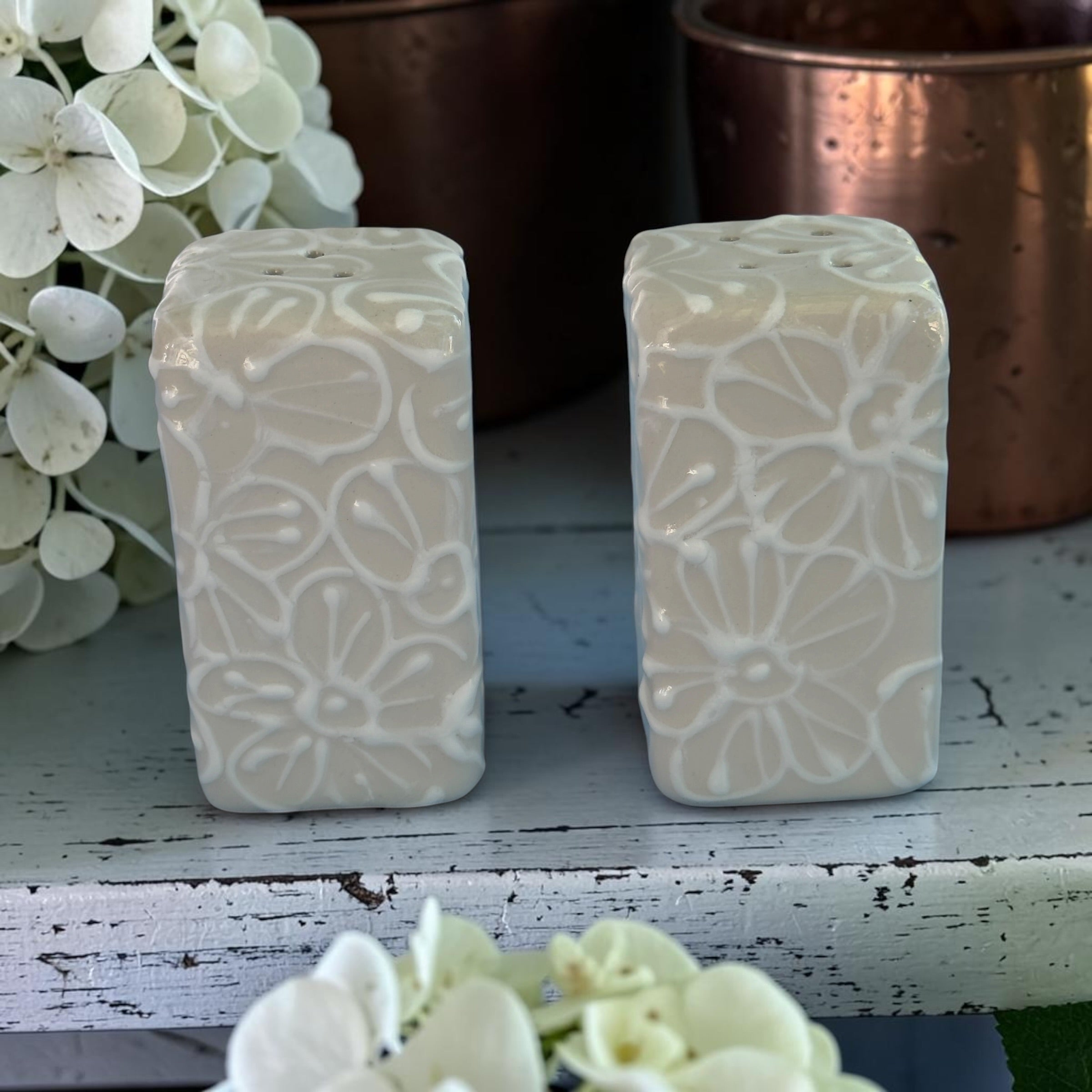 Two rectangular salt pepper ceramics  with floral patterns on a wooden surface with flowers in the background.